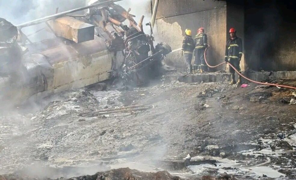 Firefighters spraying water on charred wreckage of a large vehicle.